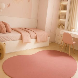 May include: A blush pink, organically shaped rug dominates the foreground of a child's bedroom. The rug's soft texture complements the room's pastel color scheme, which includes a bed with matching bedding, a desk, and a chair. The room is decorated with a sun and bird mural.