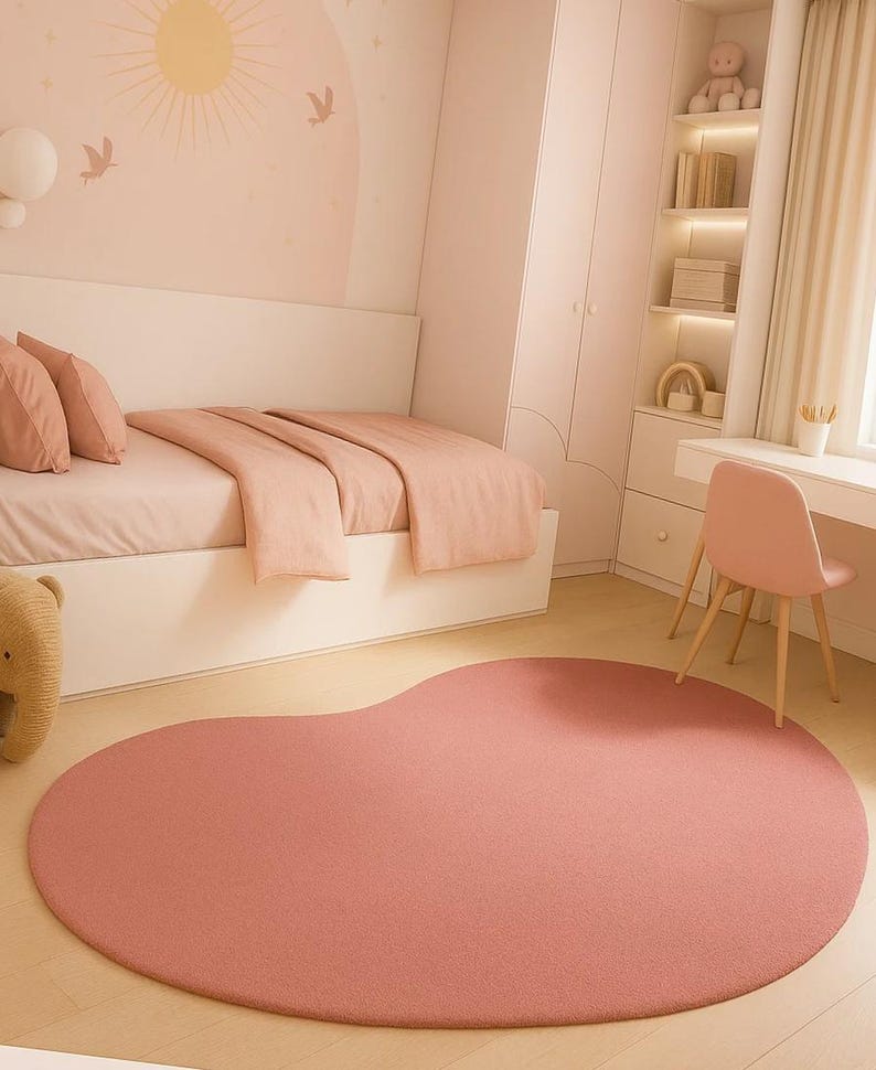 May include: A blush pink, organically shaped rug dominates the foreground of a child's bedroom. The rug's soft texture complements the room's pastel color scheme, which includes a bed with matching bedding, a desk, and a chair. The room is decorated with a sun and bird mural.