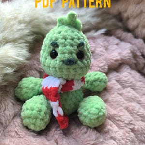 May include: Green crocheted plush toy with a red and white scarf. The toy has a grumpy expression with black eyes and a stitched-on mouth. The toy is sitting on a pink fuzzy surface.  PDF PATTERN.