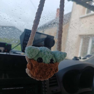 K&ouml;nnte beinhalten: Ein geh&auml;keltes Pl&uuml;schtier, das an einem R&uuml;ckspiegel in einem Auto h&auml;ngt. Das Spielzeug ist gr&uuml;n und braun und hat gro&szlig;e schwarze Augen. Das Spielzeug ist babyf&ouml;rmig.