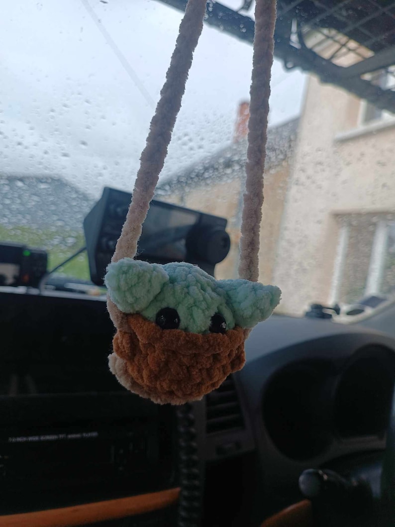 K&ouml;nnte beinhalten: Ein geh&auml;keltes Pl&uuml;schtier, das an einem R&uuml;ckspiegel in einem Auto h&auml;ngt. Das Spielzeug ist gr&uuml;n und braun und hat gro&szlig;e schwarze Augen. Das Spielzeug ist babyf&ouml;rmig.