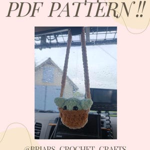 May include: A crocheted Baby Yoda hanging from a rearview mirror in a car. The crocheted figure is green and brown and is wearing a brown robe. The car is parked and the windows are wet from rain. The text "@BRIARS_CROCHET_CRAFTS" is at the bottom of the image.