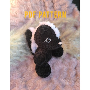 May include: A handmade crochet skunk plushie in black and white, with a fluffy white stripe down its back and a white patch on its head. The skunk has a small white nose and embroidered eyes. The text "PDF PATTERN" is in yellow.