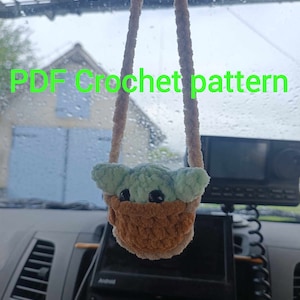 K&ouml;nnte beinhalten: Ein geh&auml;keltes Pl&uuml;schtier eines gr&uuml;nen Babycharakters, das an einem R&uuml;ckspiegel in einem Auto h&auml;ngt. Das Spielzeug tr&auml;gt einen braunen geh&auml;kelten Korb. Der Text "PDF Crochet pattern" ist im Bild sichtbar.