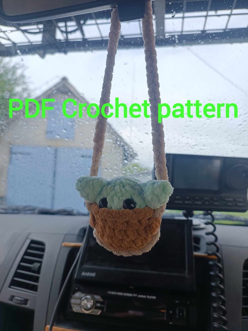 K&ouml;nnte beinhalten: Ein geh&auml;keltes Pl&uuml;schtier eines gr&uuml;nen Babycharakters, das an einem R&uuml;ckspiegel in einem Auto h&auml;ngt. Das Spielzeug tr&auml;gt einen braunen geh&auml;kelten Korb. Der Text "PDF Crochet pattern" ist im Bild sichtbar.