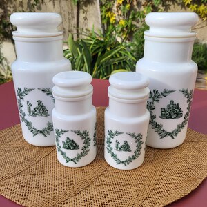 Peut inclure: Ensemble de quatre pots à épices en céramique blanche avec des motifs floraux verts et blancs. Les pots ont des couvercles et sont disposés sur un dessous de plat en osier brun.