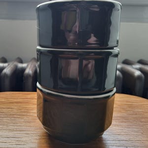 Puede incluir: Un juego de tres tazas de cerámica de color verde oscuro apiladas una encima de la otra. Las tazas tienen una superficie ligeramente texturizada y una forma redondeada.