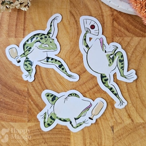 May include: Three whimsical frog stickers in various poses. The frogs are white with green spots and black outlines. One frog holds a fan, another a bottle, and the third is mid-movement. The stickers are on a wooden surface.