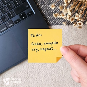 Puede incluir: Una nota adhesiva amarilla con el texto "To do: Code, compile, cry, repeat..." se sostiene cerca del teclado de un portátil. La nota está enfocada, con el portátil y un fondo suave y estampado ligeramente borrosos.