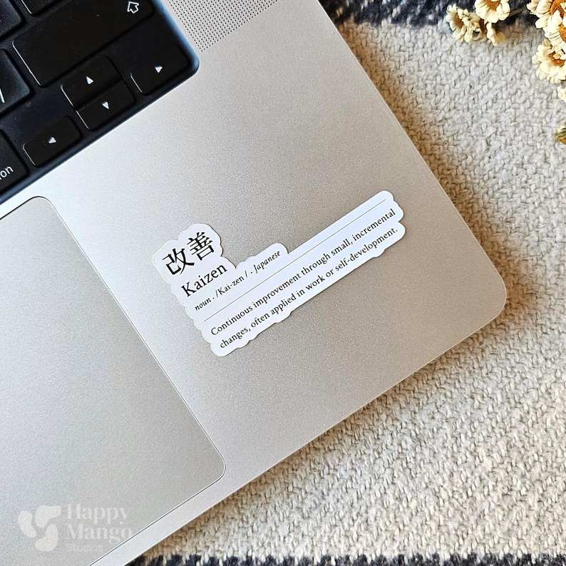May include: A white sticker with black text on a silver laptop. The sticker says "改善 Kaizen noun /Kai-zen/ Japanese Continuous improvement through small, incremental changes, often applied in work or self-development."