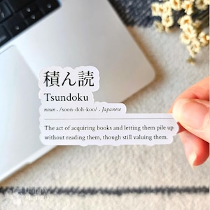 May include: A white sticker with black text defines the Japanese word "Tsundoku" as the act of acquiring books and letting them pile up without reading them, though still valuing them.