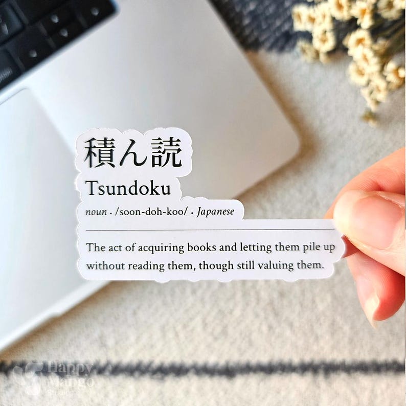 May include: A white sticker with black text defines the Japanese word "Tsundoku" as the act of acquiring books and letting them pile up without reading them, though still valuing them.