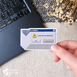 Pode incluir: Uma pilha de adesivos brancos com um design retrô de mensagem de erro de computador. A mensagem diz "WARNING! Lorem ipsum detected." com um ponto de exclamação e um símbolo de aviso triangular amarelo. Os adesivos são segurados em uma mão.