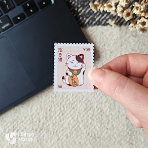 Op de afbeelding: Een postzegel met een cartoon Maneki-neko kat met een opgeheven poot, die een gouden munt vasthoudt. De postzegel heeft Japanse tekst en een waarde van ¥50. De kat heeft een wit lichaam, zwarte en bruine markeringen en een groene kraag.