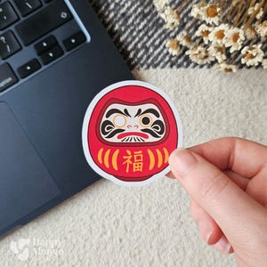 May include: A red Daruma doll sticker with black and yellow facial features and Japanese text. The sticker is round and held by a hand, with a laptop and flowers in the background.