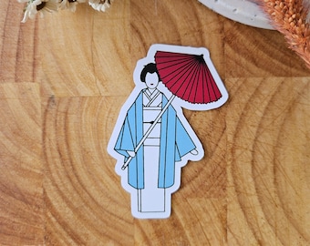 May include: A sticker featuring a Japanese geisha in a kimono, holding a red parasol. The geisha is wearing a light blue kimono with white and black accents. The sticker has a white border and is on a wooden surface.