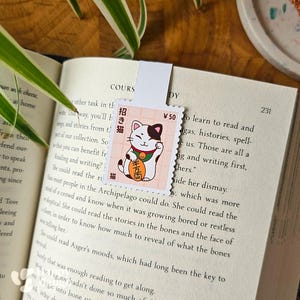 May include: Bookmark with a stamp design featuring a Japanese Maneki-neko cat illustration. The cat is white with black markings, holding a gold coin. The bookmark is in an open book.