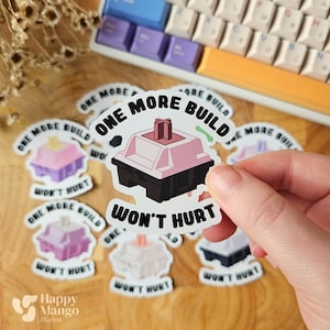 Op de afbeelding: Een verzameling stickers met een mechanisch keyboardschakelaarontwerp. De stickers hebben een roze, zwarte en witte kleurstelling, met de tekst "ONE MORE BUILD WON'T HURT". Een toetsenbord is zichtbaar op de achtergrond.