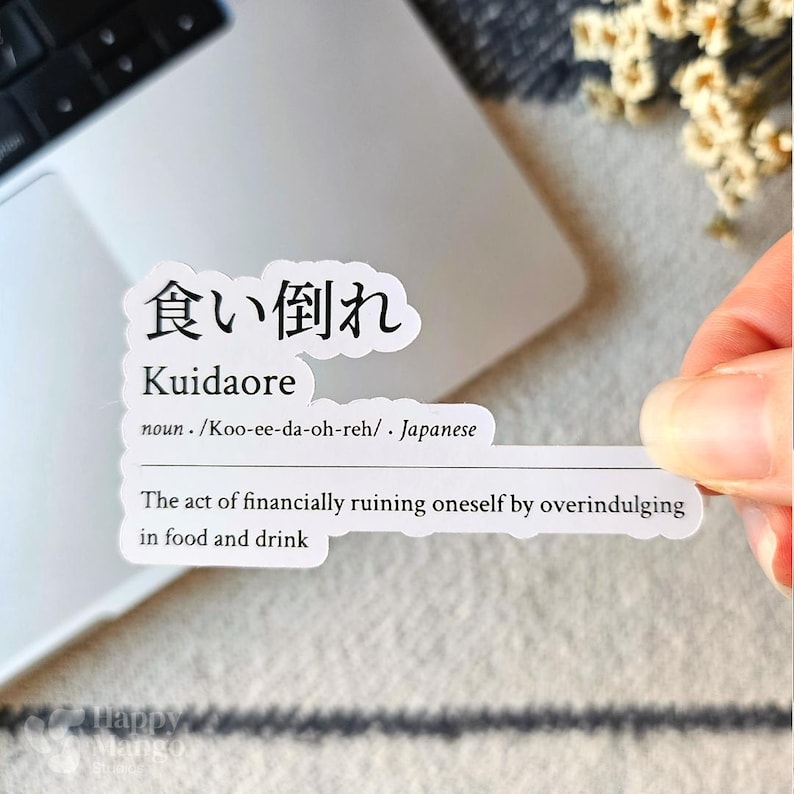 May include: A white sticker with black text defines the Japanese word "Kuidaore" as the act of financially ruining oneself by overindulging in food and drink. The word is written in Japanese characters and the definition is in English.