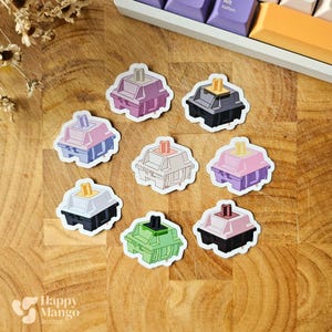 May include: A collection of colorful mechanical keyboard switch stickers in various colors, including pink, purple, black, and green. The stickers are arranged on a wooden surface, with a keyboard in the background. The Happy Mango Studios logo is visible.
