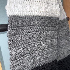 May include: A handmade crochet scarf in a color block design. The scarf features sections of white, gray, and black. The crochet stitch creates a textured pattern, adding visual interest to the accessory. The scarf is draped over a dark metal rod.