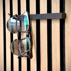 Könnte beinhalten: Ein schwarzer Metall-Brillenhalter an einer Holzwand mit vertikalen Streifen. Zwei Brillenpaare hängen an dem Halter. Eine hat einen Schildpattrahmen, die andere einen silbernen Rahmen.