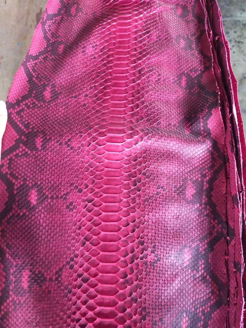 May include: Close-up of a stack of pink snakeskin leather. The leather has a scale pattern with dark brown markings. The central spine of the snakeskin is a solid pink color. The leather is likely used for fashion accessories or upholstery.