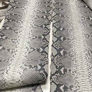 以下が含まれることがあります： グレーと白のヘビ革のクローズアップ。革は、グレーと白のトーンのバリエーションを持つ鱗模様です。画像には、ヘビ革のいくつかの部分が写っており、一部は折りたたまれています。