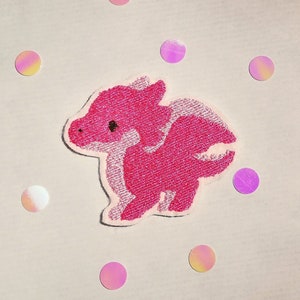 Könnte beinhalten: Rosa gestickter Bügelaufkleber mit einem Cartoon-Drachen mit weißem Umriss.