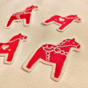Könnte beinhalten: Vier gestickte Dala-Pferde-Patches in Rot und Weiß. Die Pferde sind traditionelle schwedische Volkskunst.