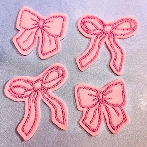 Może przedstawiać: Cztery różowe wstążki z filcu z różowym haftowanym obramowaniem. Wstążki są ułożone w kwadratowy wzór na jasnoniebieskim tle.