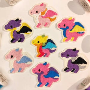 May include: Embroidered dragon patches in various colors, including pink, purple, yellow, and blue. Each patch features a unique dragon design with detailed stitching and a white border. Spools of thread in various colors are scattered around the patches.