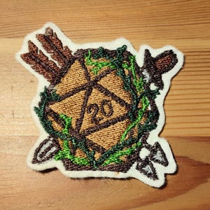 Op de afbeelding: Geborduurde patch met een bruine D20 dobbelsteen met het nummer 20, omringd door groene wijnranken en pijlen. De patch heeft een witte rand en is ontworpen voor gaming liefhebbers. Het is waarschijnlijk bedoeld voor kleding of tassen.