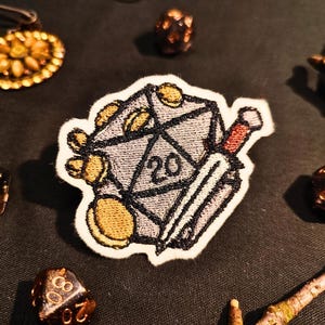 Op de afbeelding: Gestikt patch met een 20-zijdige dobbelsteen met een zwaard en gouden munten. De dobbelsteen is grijs met zwarte contouren en het getal 20 is in zwart gedrukt. Het zwaard is zilverkleurig met een rode handgreep. De munten zijn goudkleurig met zwarte contouren.