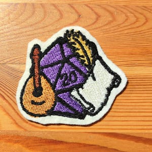 Könnte beinhalten: Gestickter Aufnäher mit einer Mandoline, einem lila zwanzigseitigen Würfel, einer Feder und einer Schriftrolle. Der Aufnäher hat einen weißen Rand und ist vor einem Holzgrund platziert. Das Design bezieht sich wahrscheinlich auf Rollenspiele.