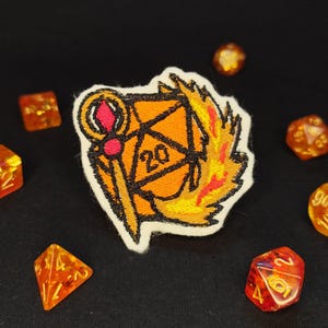Op de afbeelding: Een geborduurde patch met een oranje twintigzijdige dobbelsteen met het getal 20, een staf met een roze edelsteen en gele en oranje vlammen. De patch is omringd door verschillende doorschijnende oranje en rode veelzijdige dobbelstenen op zwart.