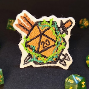 Op de afbeelding: Een geborduurde patch met een oranje twintigzijdige dobbelsteen met het zwarte cijfer 20. De dobbelsteen is omstrengeld met felgroene ranken en doorboord door twee gekruiste pijlen met oranje bevedering en grijze punten op een witte stoffen ondergrond.
