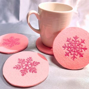 Könnte beinhalten: Vier runde Untersetzer aus rosa Filz mit rosa gestickten Schneeflocken. Die Untersetzer sind rund und auf einem liegt eine weiße Tasse.