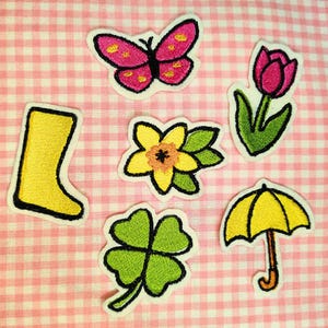 May include: A set of six embroidered iron-on patches featuring a yellow rain boot, a pink butterfly, a yellow umbrella, a green four-leaf clover, a yellow daffodil, and a pink tulip.