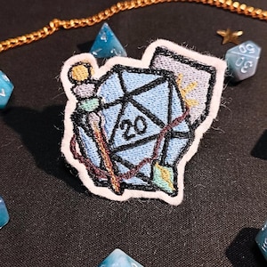 May include: A blue and white embroidered patch featuring a twenty-sided die, a wand, and a rolled-up scroll. The patch is surrounded by a white border.