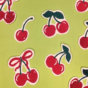 Könnte beinhalten: Gestickte Kirsch-Patches auf hellgrünem Hintergrund. Die Kirschen sind rot mit grünen Blättern und Stielen, einige mit roter Schleife. Die Patches haben einen weißen Rand.
