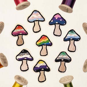 Può includere: Otto toppe ricamate a forma di fungo disposte in cerchio, ognuna con un motivo di colore unico sul cappello e un gambo beige. Le toppe sono circondate da otto rocchetti di filo da cucito colorati in rosso, viola, blu, giallo e verde.
