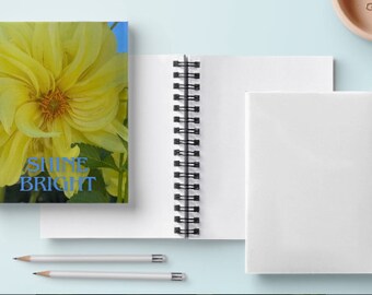 May include: A spiral bound notebook with a yellow flower on the front cover. The flower is a dahlia with a yellow center and white petals. The text "Shine Bright" is printed in blue letters on the cover.
