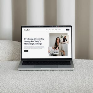 May include: A silver laptop displaying a website with the text "Developing A Compelling Strategy For Today's Marketing Landscape." The website features images of a woman in a blazer and a woman working at a table. The laptop is on a white, textured surface.