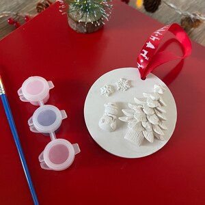 Puede incluir: Adorno navideño redondo blanco con diseño de muñeco de nieve y árbol de Navidad. Tiene una cinta roja para colgar. Incluye tres pequeños botes de pintura rosa, azul y roja, y un pincel azul. El fondo es una superficie roja.