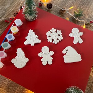 May include: A set of six white ceramic Christmas ornaments, including a snowman, a gingerbread person, a Christmas tree, a snowflake, a Santa hat, and a second gingerbread person. The ornaments are on a red background with paint pots and a paintbrush.