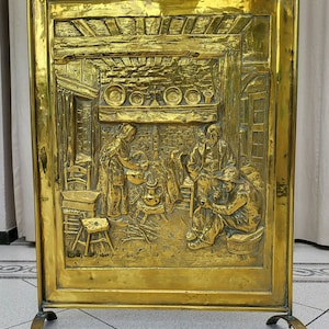 Peut inclure: Un pare-feu en laiton avec un motif en relief représentant une scène de personnes dans une pièce. L'écran a une poignée en anneau en haut et trois pieds.