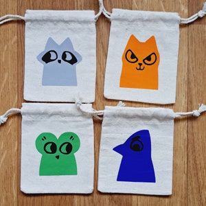 May include: Four small white fabric bags with drawstring closures. Each bag features a different cartoon animal: a raccoon, a cat, a frog, and a bird. The animals are drawn in a simple, playful style and are coloured in bright, bold colours.