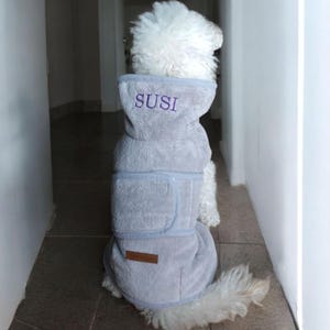 Può includere: Un cane bianco con un cappotto con cappuccio grigio. Il cappotto ha un bordo blu chiaro e il nome "SUSI" ricamato sul cappuccio in viola. Una piccola toppa marrone è sulla parte inferiore del cappotto.