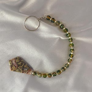 May include: A keychain with a patterned stone pendant and a beaded strand. The beads are green and gold, and the keychain has a silver-coloured key ring. The pendant is a mix of colours.